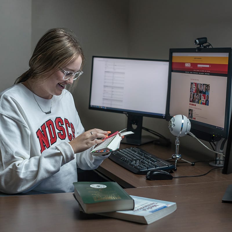 Smiling student in an NDSCS sweatshirt flipping through a notebook at a desk with dual monitors and a microphone.