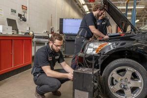 Three male students in NDSCS uniforms diagnosing a black car with the hood open in an automotive lab, using diagnostic equipment and a connected computer.