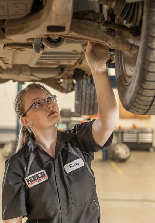 Female automotive student wearing NDSCS uniform inspecting the underside of a vehicle in a garage setting.