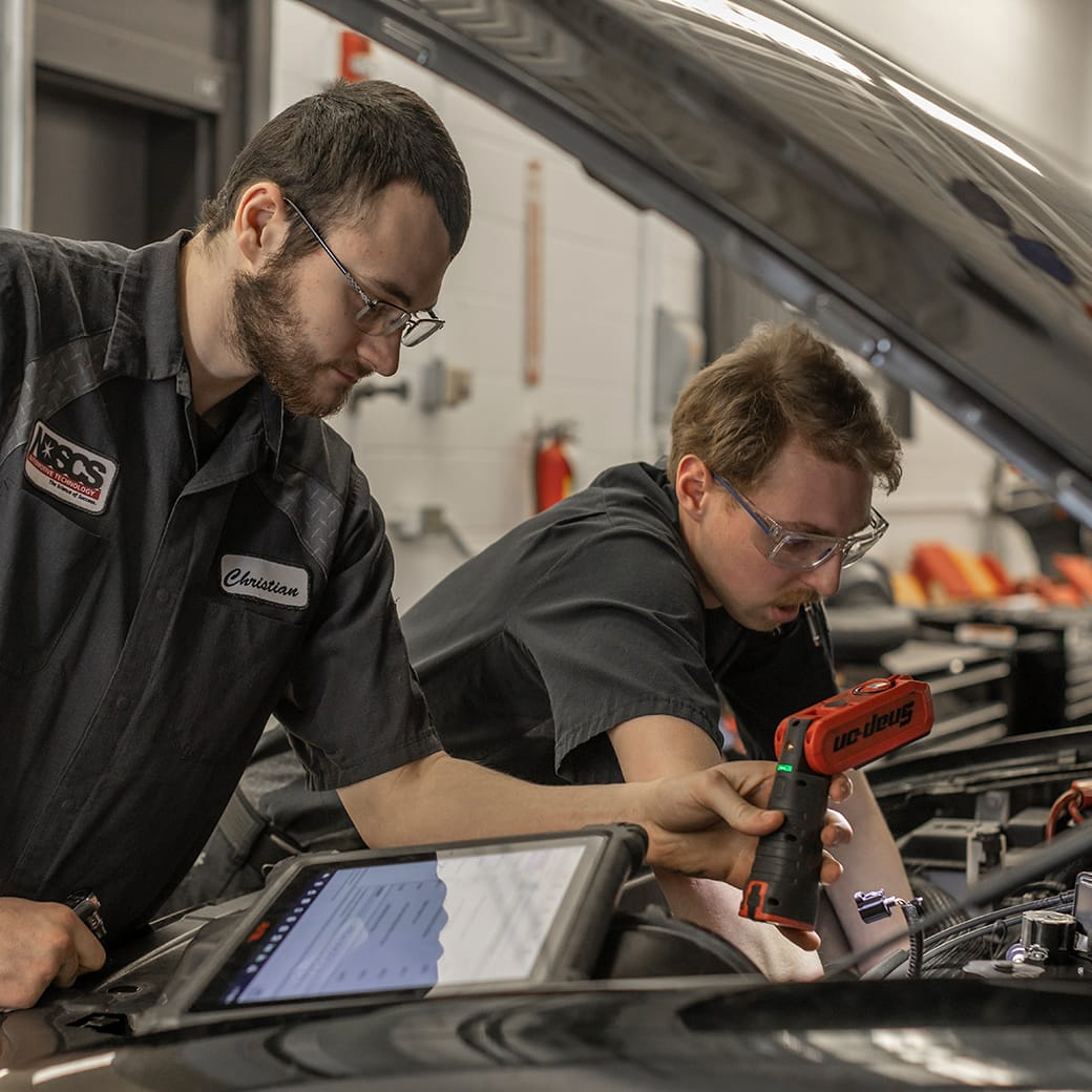 Two automotive students wearing safety glasses examining an engine, one holding a diagnostic tool and the other reviewing information on a tablet.