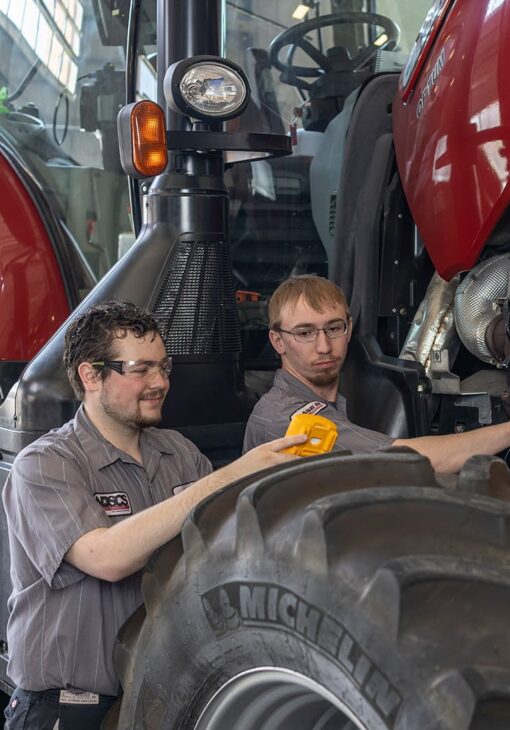 Two male students in NDSCS uniforms using a diagnostic tool on a red Case IH tractor, with one student pointing the tool and both wearing safety glasses.
