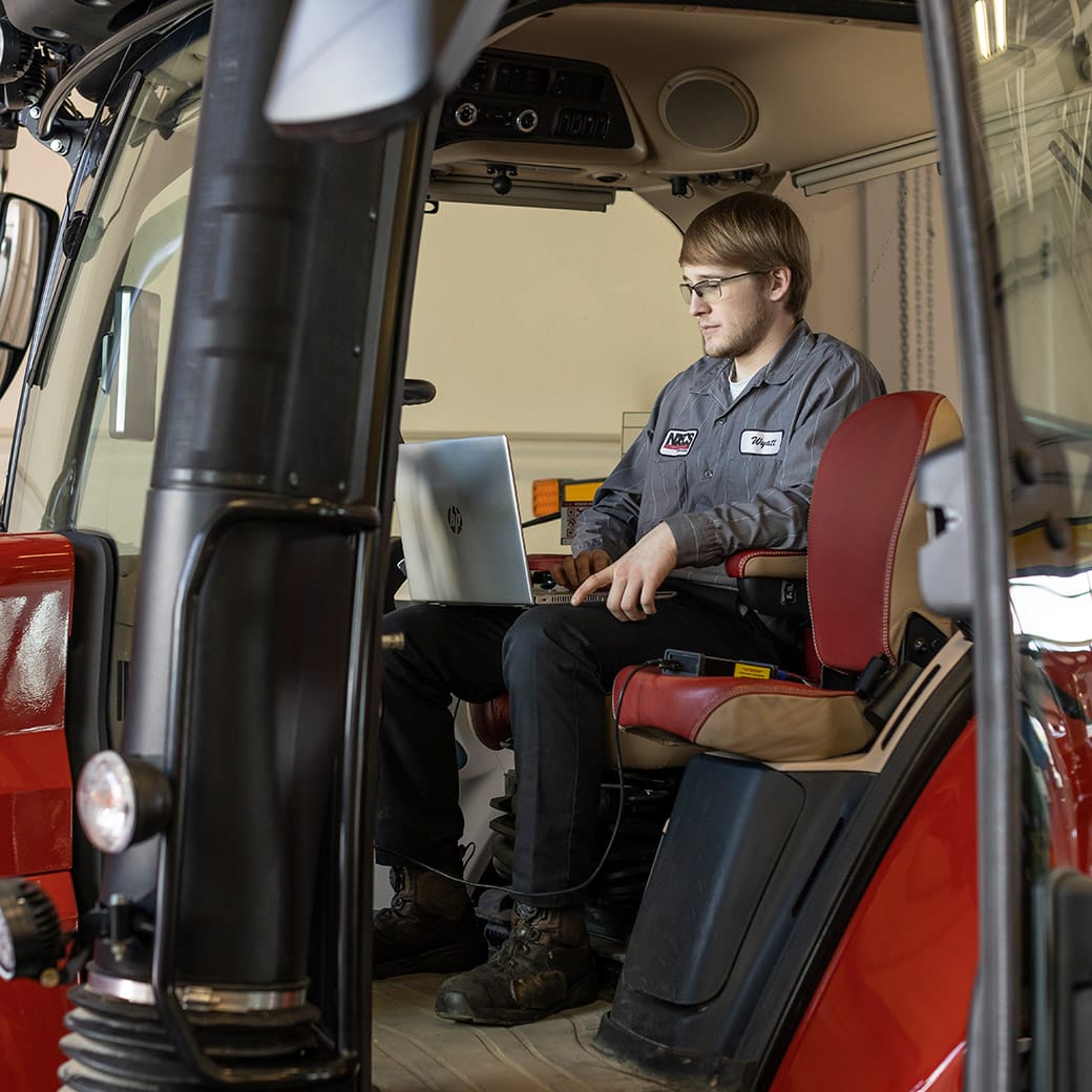 Male student sitting inside a red tractor cab using a laptop for diagnostics, wearing safety glasses and an NDSCS uniform.