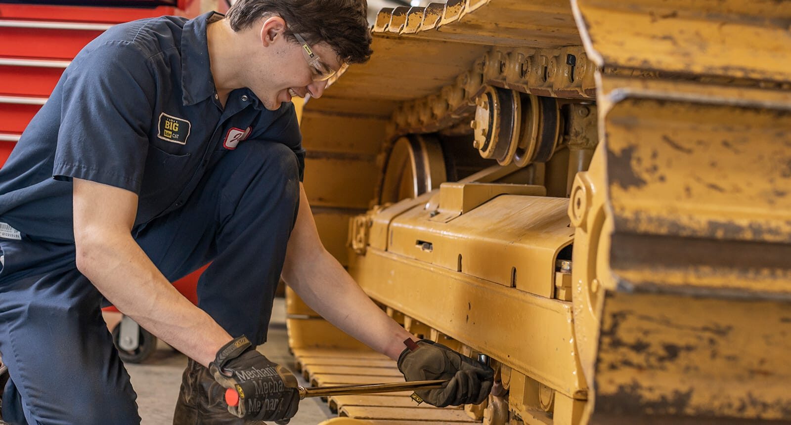 Mechanic in navy coveralls using a wrench to work on the track of a large yellow construction vehicle inside a garage.