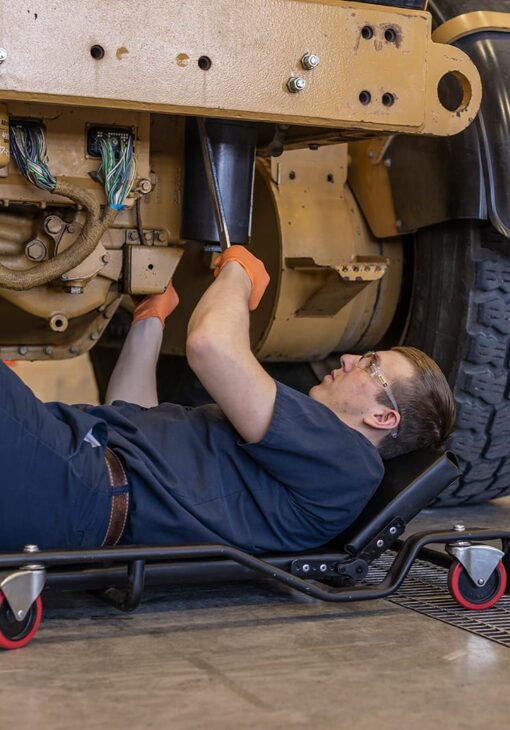 Mechanic lying on a creeper beneath a large vehicle, using a tool to work on the undercarriage while wearing safety glasses and orange gloves.