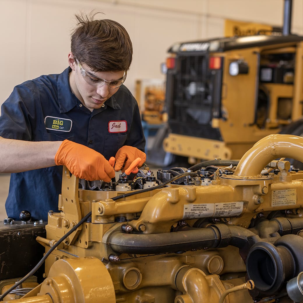 Mechanic in safety glasses and orange gloves working on the engine of heavy equipment inside a shop, with a yellow bulldozer in the background.