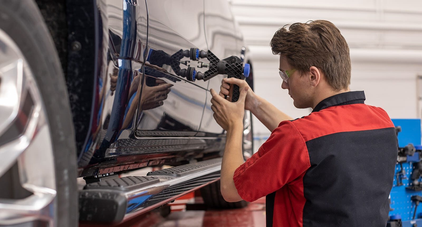 Male student in a red and black uniform using a paintless dent repair tool on the side panel of a black vehicle inside an auto body shop.