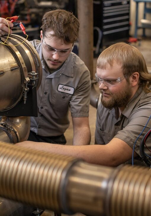 Two students in safety glasses working together on a large industrial machine in a workshop.