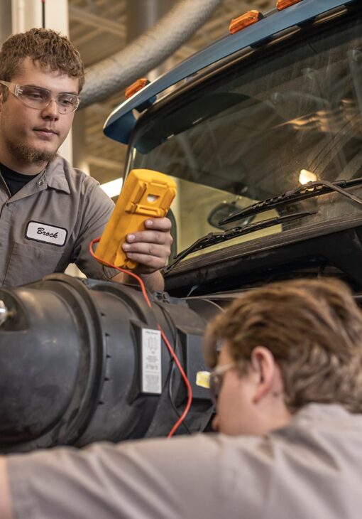 Two male students in NDSCS diesel uniforms using a multimeter to diagnose a large truck engine in a workshop.