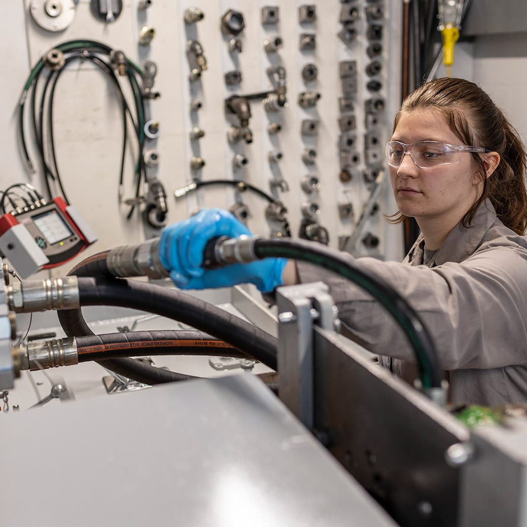 Female student wearing safety glasses and gloves connecting hydraulic hoses in a lab with metal fittings organized on the wall behind her.
