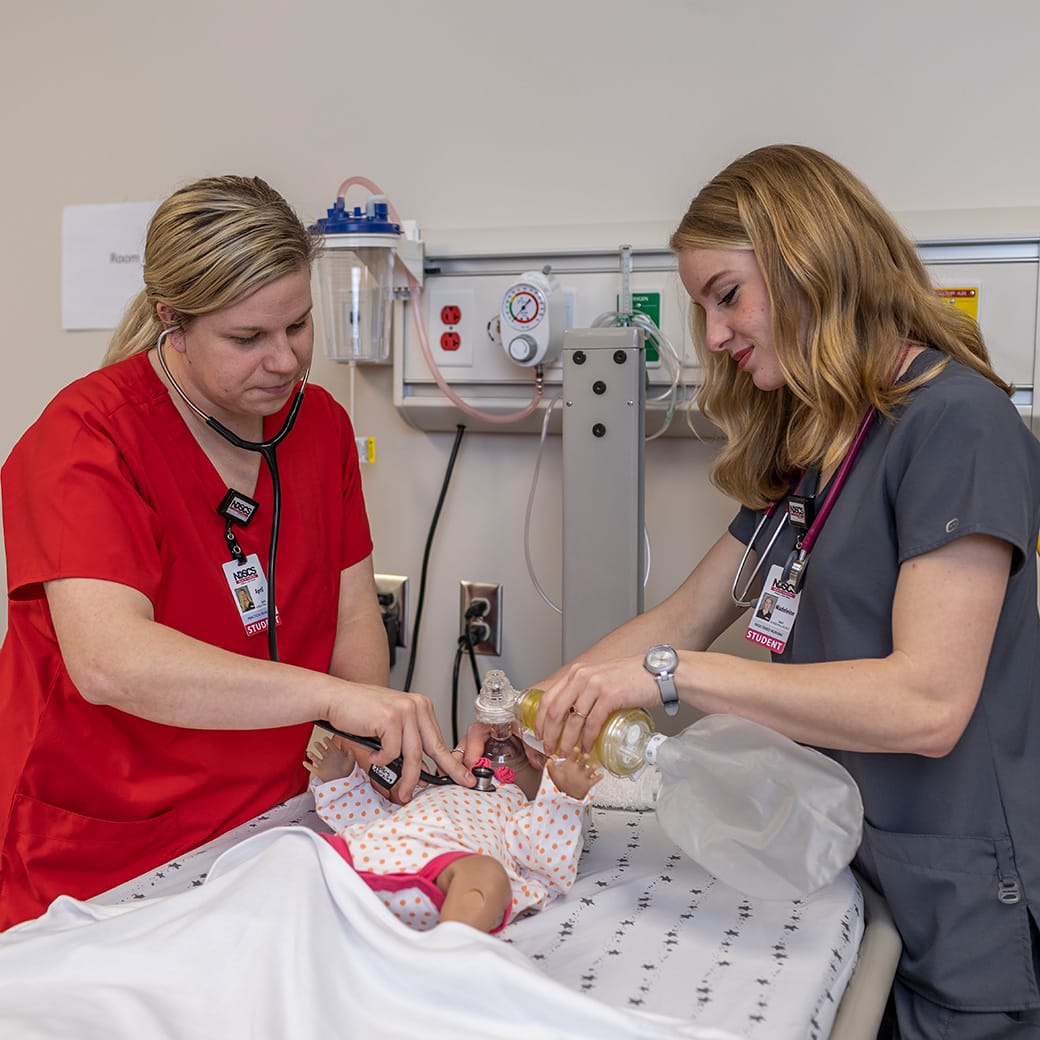 Two nursing students performing a simulated infant emergency procedure using a training mannequin in a clinical lab setting.
