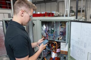 Student in safety glasses holds a tablet while inspecting the interior wiring of an open HVAC unit in a classroom workshop.