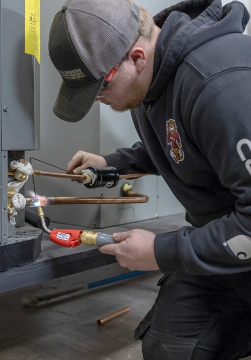 Student in a hoodie and safety glasses uses a torch to solder copper piping on an HVAC unit in a workshop setting.