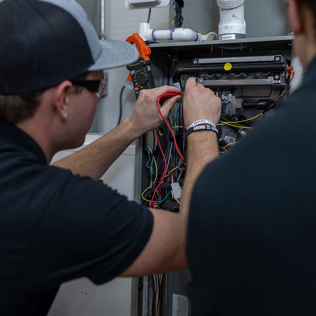 Two students in black shirts and safety glasses work together on an open HVAC unit, using multimeter probes to test wiring connections.