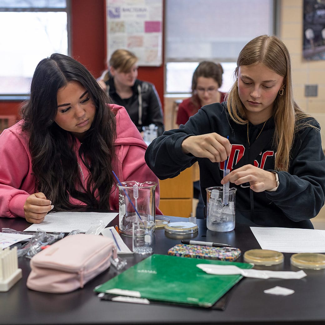 Two female students working together in a science lab, one reading instructions while the other places a test tube into a beaker filled with ice.