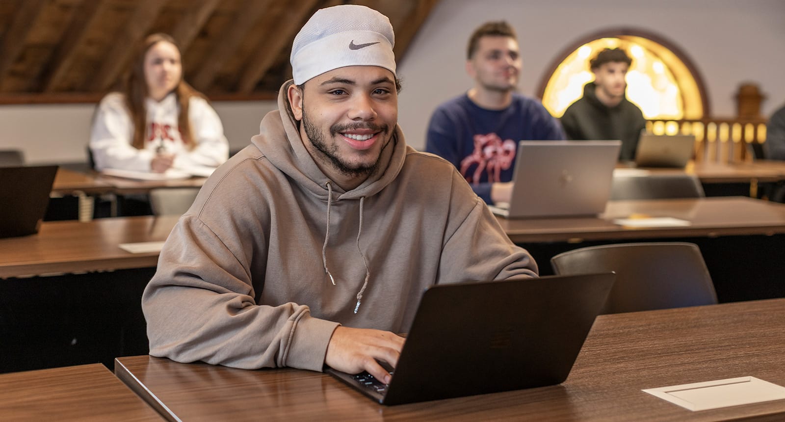 Smiling student in a hoodie working on a laptop in a classroom with other students in the background
