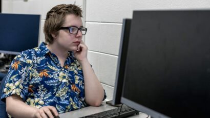Student in a floral shirt focused on a computer screen in a classroom with white brick walls.