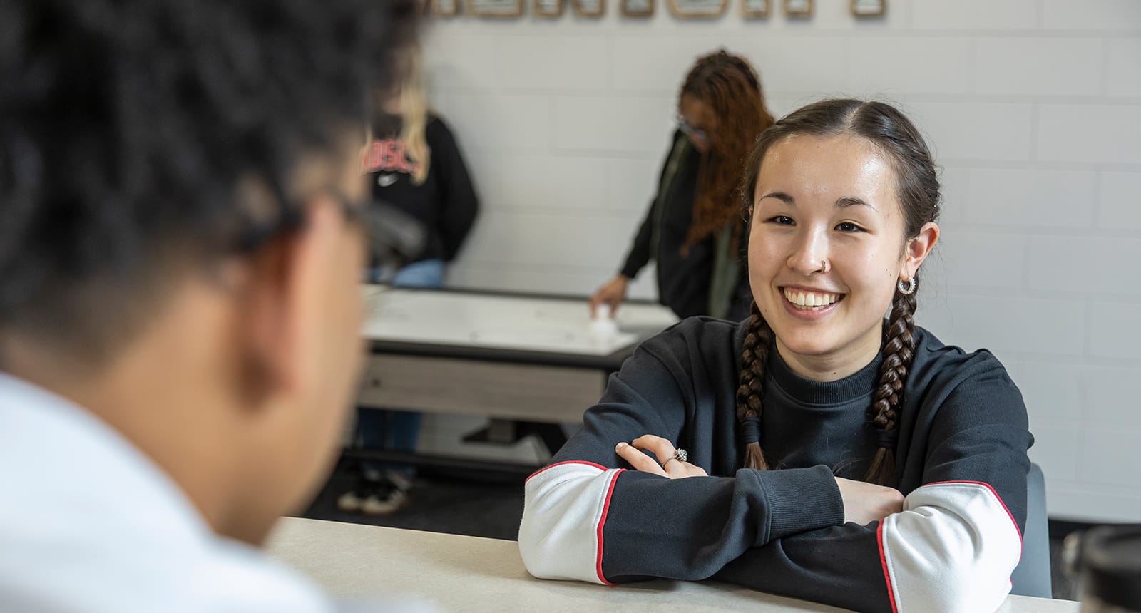 Smiling student with braided hair sits at a table talking to a peer, with other students playing air hockey in the background.