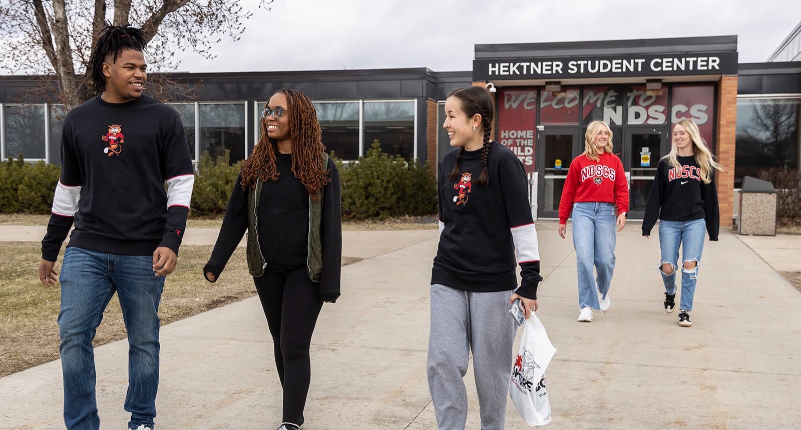 Group of six college students walking and talking outside the Hektner Student Center at NDSCS, wearing branded sweatshirts and casual clothing.