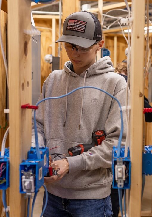 Student in safety glasses and a baseball cap wiring blue electrical boxes in a wooden training frame, with a power drill tucked under their arm.
