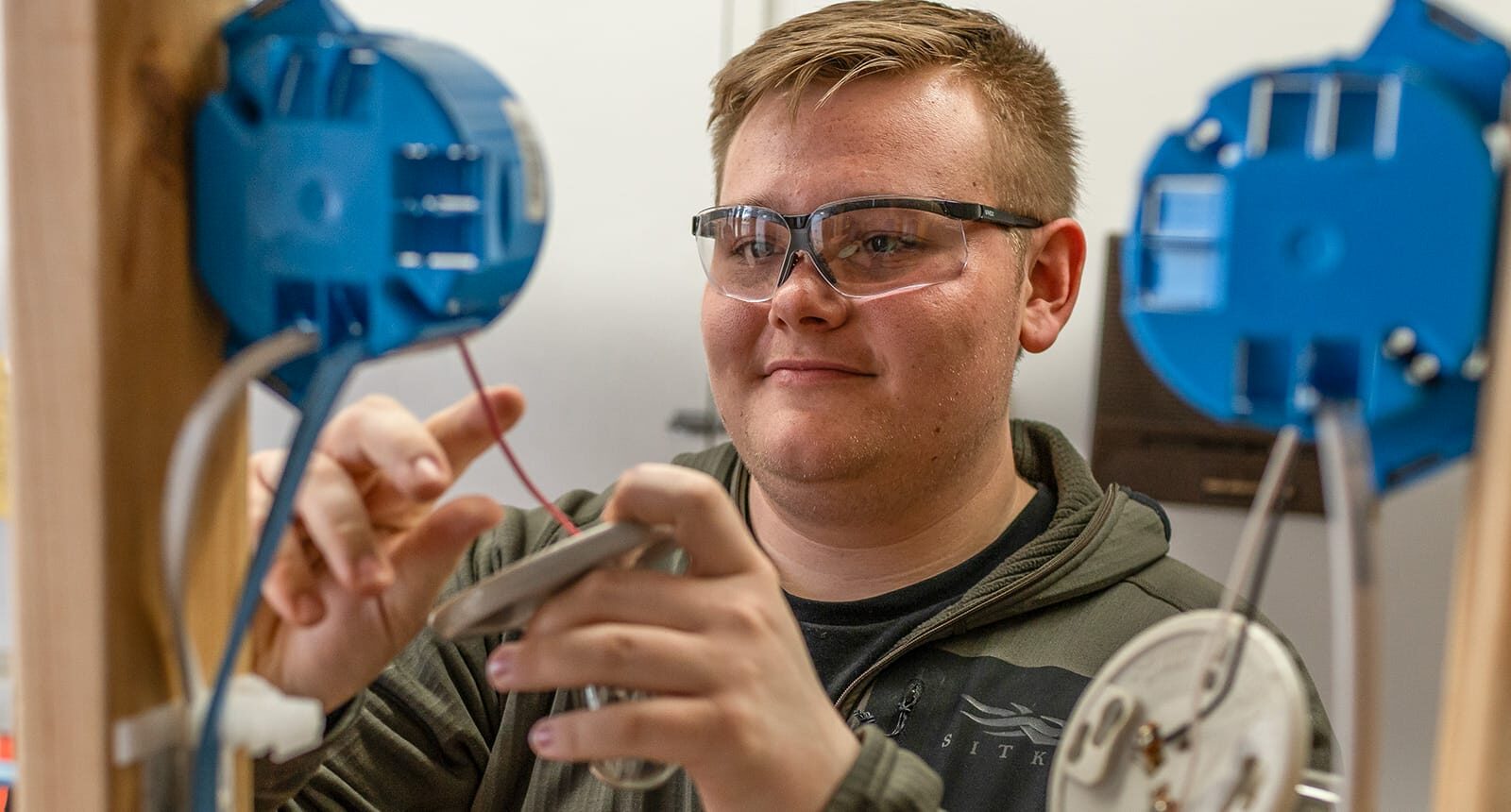 Smiling student wearing safety glasses wiring an electrical outlet during hands-on training, with blue outlet boxes mounted on wooden frames.