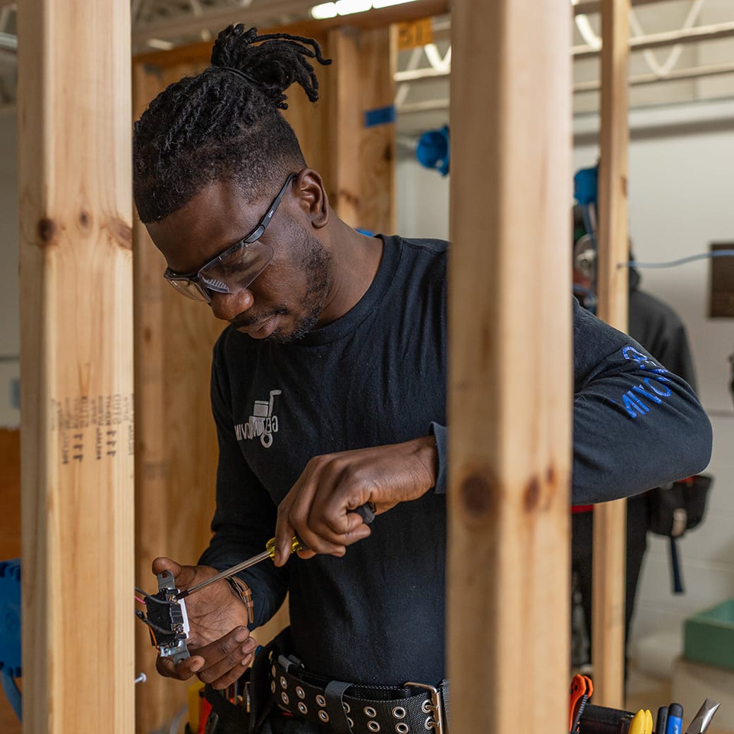 Student wearing safety glasses and a tool belt wiring an electrical outlet between wooden framing studs during hands-on training.