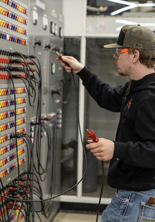 Student wearing safety glasses and a cap working with test cables on a large electrical control panel filled with multicolored connection ports.