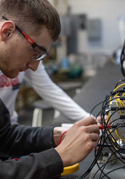 Student wearing safety glasses working with multicolored wires on an electrical testing panel in a lab environment.