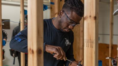 Student wearing safety glasses uses a screwdriver to install electrical wiring between wooden framing beams.