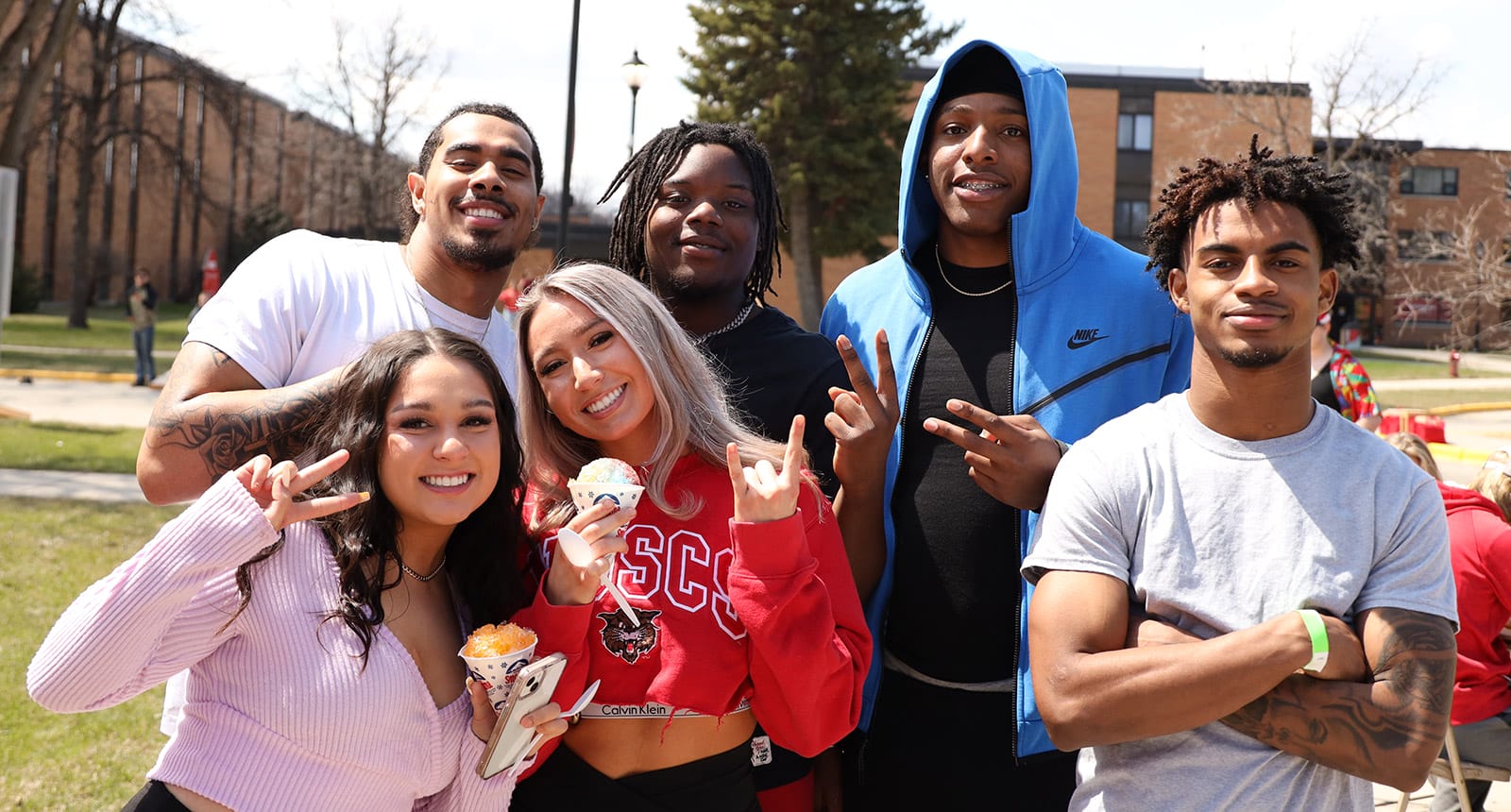 Group of six college students posing and smiling outdoors on campus during a sunny day, with two holding snow cones and others flashing peace signs and hand gestures.