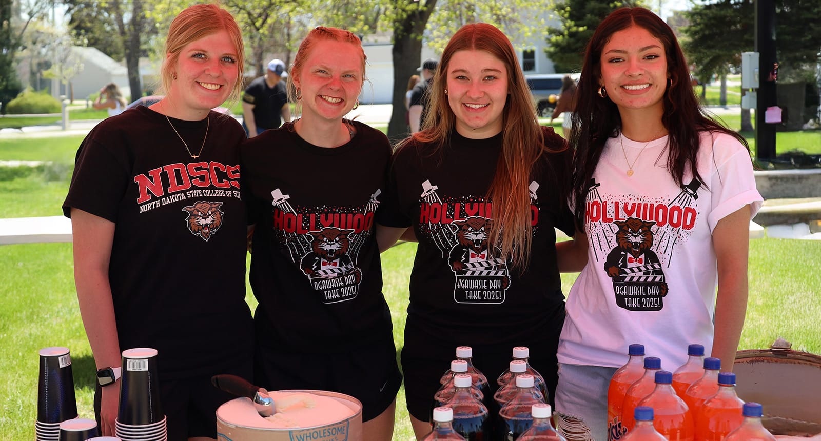 Four young women standing behind a table with drinks and cups, smiling and wearing NDSCS or “Hollywood Agawasie Day Take 2025!” T-shirts, outdoors on a sunny day.
