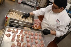 Culinary student in white chef coat wrapping bacon around scallops on a metal tray in a commercial kitchen.