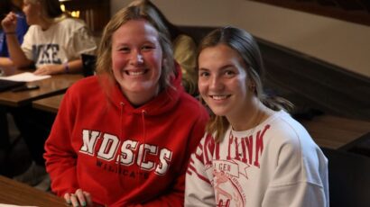 Two students smiling at a table in a classroom or study space, wearing NDSCS and GEM-themed sweatshirts.