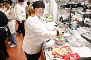 Student in a white chef’s coat preparing food at a counter with pita bread and mixing bowls, while other students work in the background of a busy kitchen.