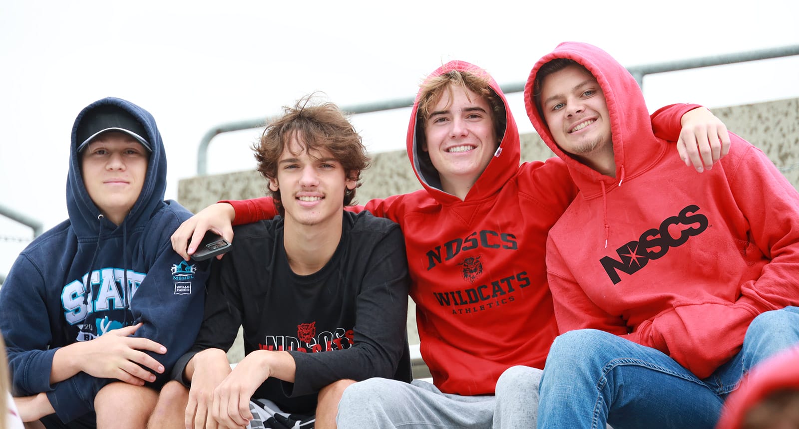 Four young men sitting together outdoors, smiling, wearing hoodies and sweatshirts with NDSCS and sports team logos.