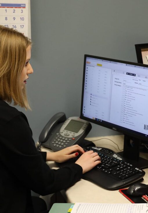 Student working at a desk with a desktop computer, typing with both hands while reviewing a spreadsheet or checklist on screen, with a phone and calendar in the background.