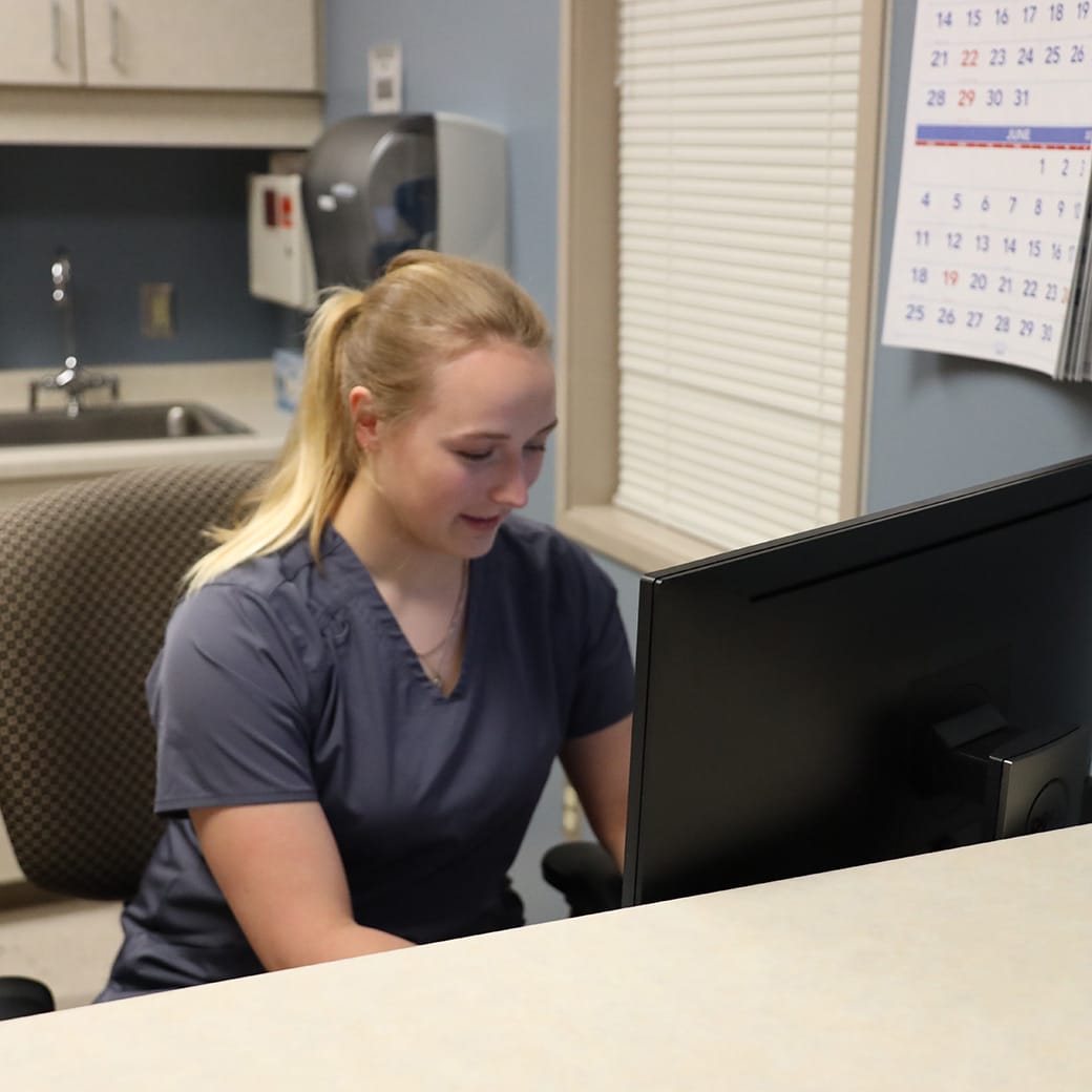 Student in scrubs working at a computer station in a medical office with a calendar on the wall and a sink in the background.