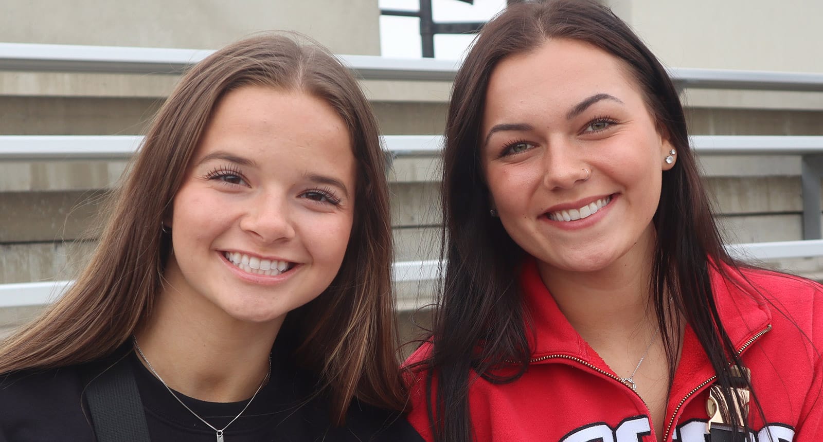 Two young women smiling while sitting on bleachers, one wearing a black shirt and the other wearing a red NDSCS jacket.