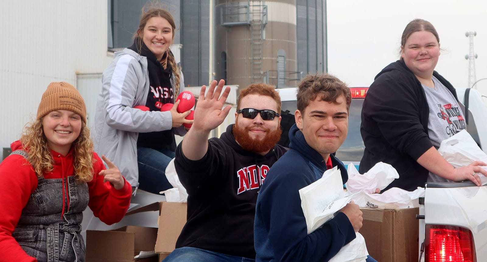Group of five students sitting on a pickup truck, smiling and holding bags and items for distribution.