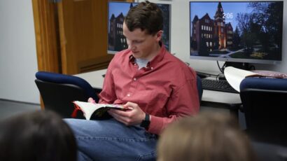 Man in a red shirt reading aloud from a book to a small group in a computer lab.