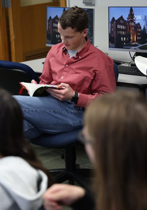 Man in a red button-up shirt reading a book while seated in a computer lab, with students in the foreground.