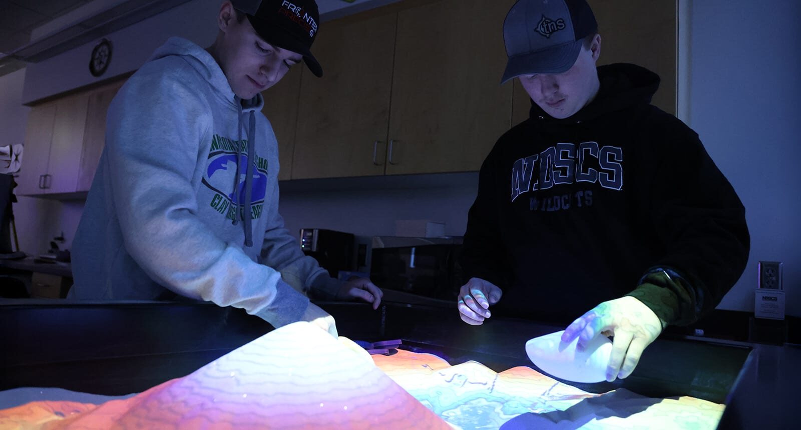 Two students in hoodies and hats use a 3D augmented reality sandbox under dark lighting in a classroom lab.