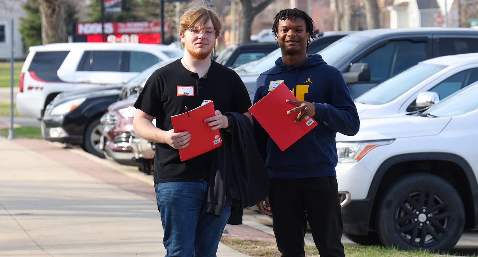 Two male students walking outside on campus holding red folders, with parked cars in the background.