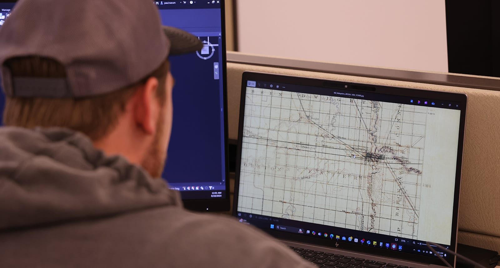 Student wearing a gray hoodie and cap working at a desk with dual monitors, one displaying a historical map on a laptop screen.