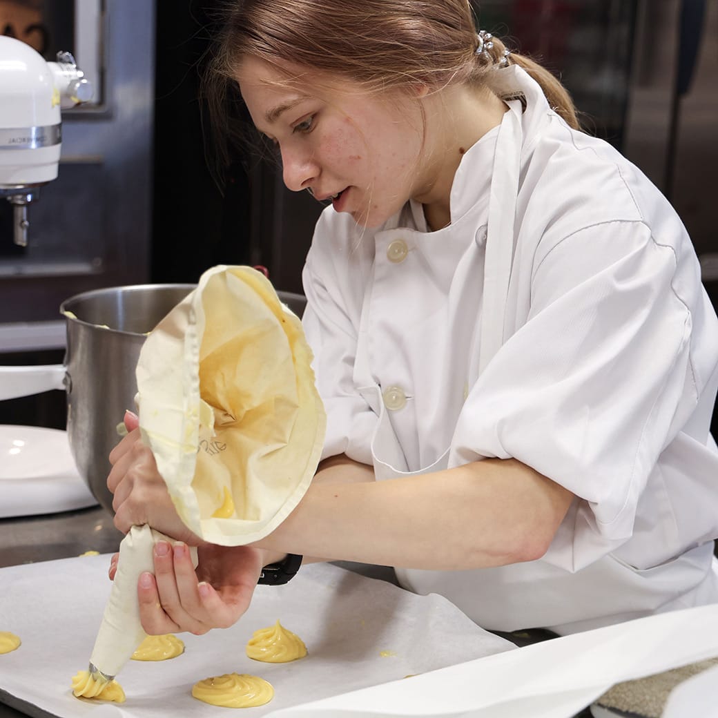 Culinary student in a white chef coat piping dough onto a parchment-lined baking sheet in a commercial kitchen.