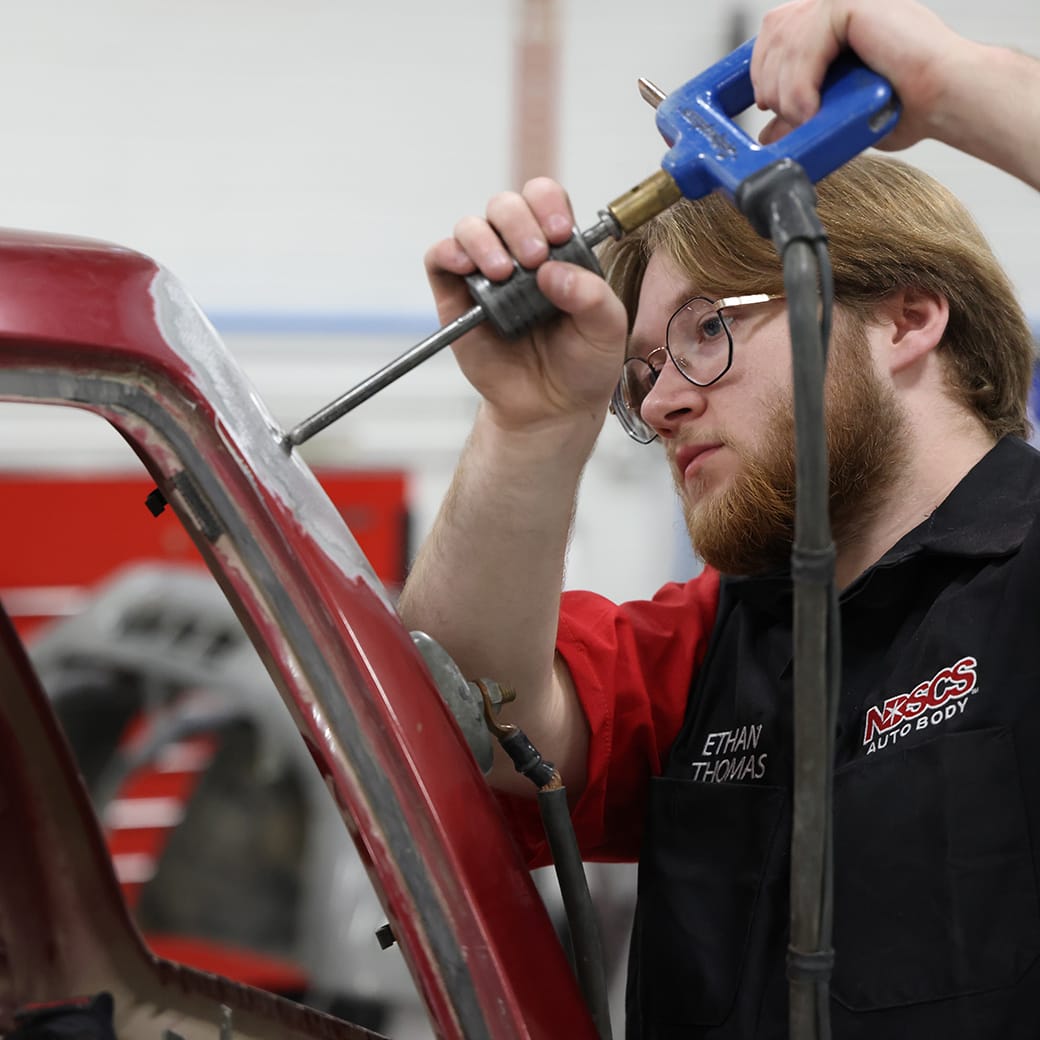 Male student wearing safety glasses and an NDSCS Auto Body uniform using a pneumatic tool to repair a red vehicle frame.
