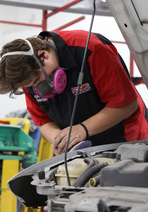 Student in an NDSCS Auto Body uniform wearing a respirator and sanding a car part under the open hood of a vehicle.