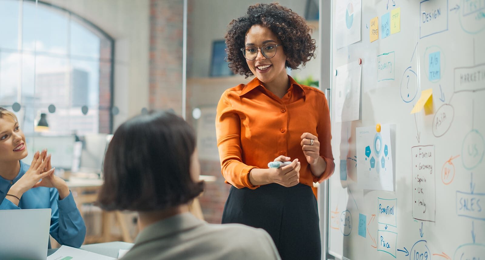 Smiling woman in orange blouse leading a strategy meeting, presenting ideas on a whiteboard covered in charts and sticky notes, with two colleagues listening attentively.