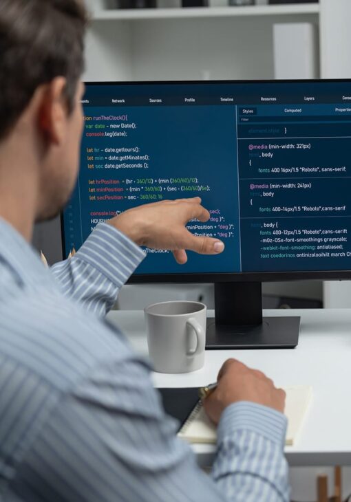 Man pointing at lines of code on a computer monitor while writing in a notebook at a desk.