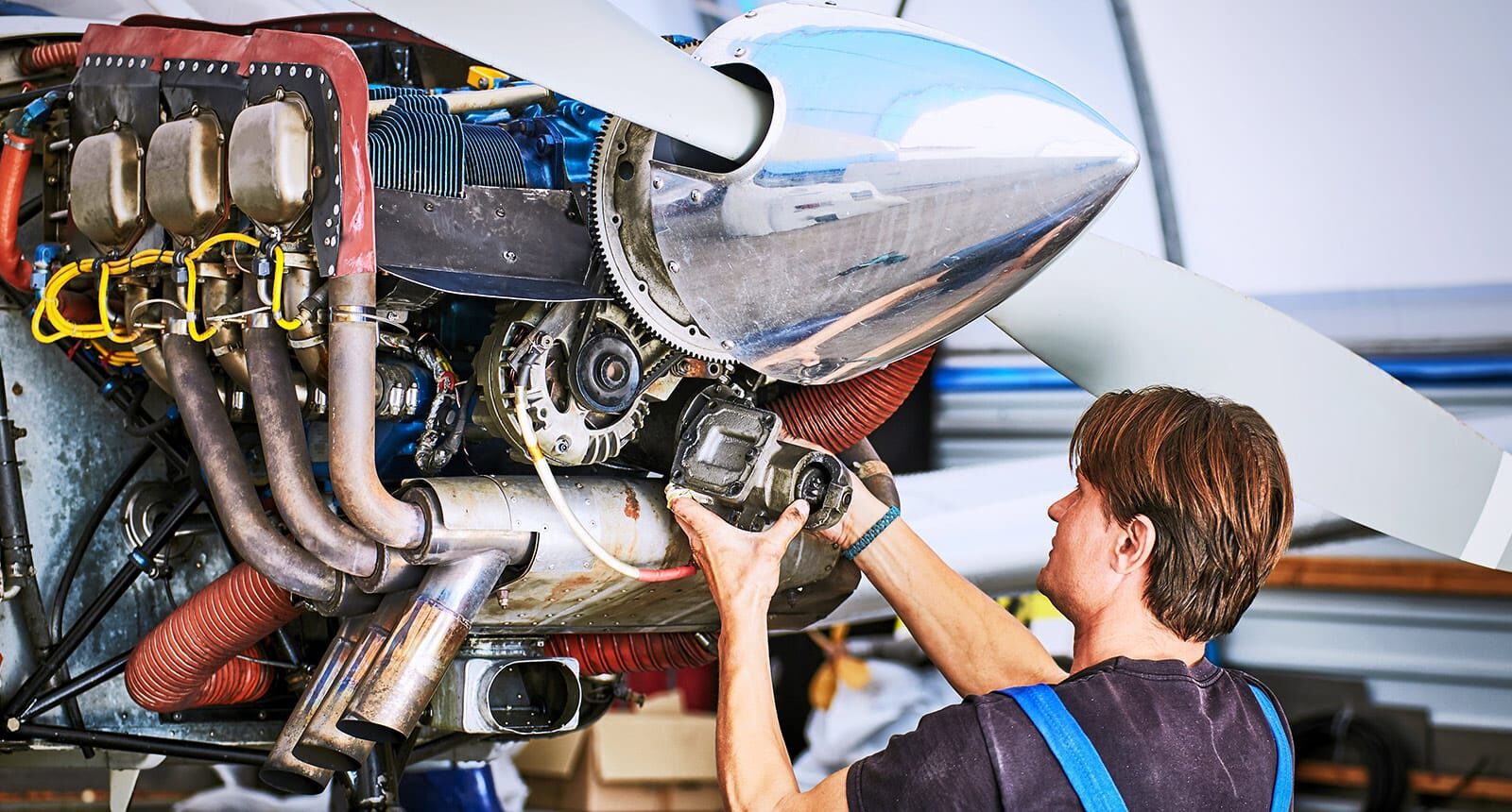 Technician working on the engine of a small aircraft with exposed parts and large propeller in a hangar.