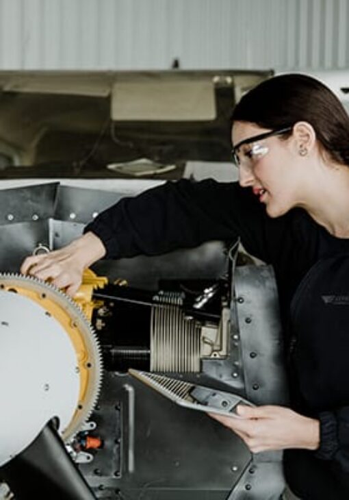 Female aviation technician inspecting a small aircraft engine while holding a clipboard, inside a hangar.